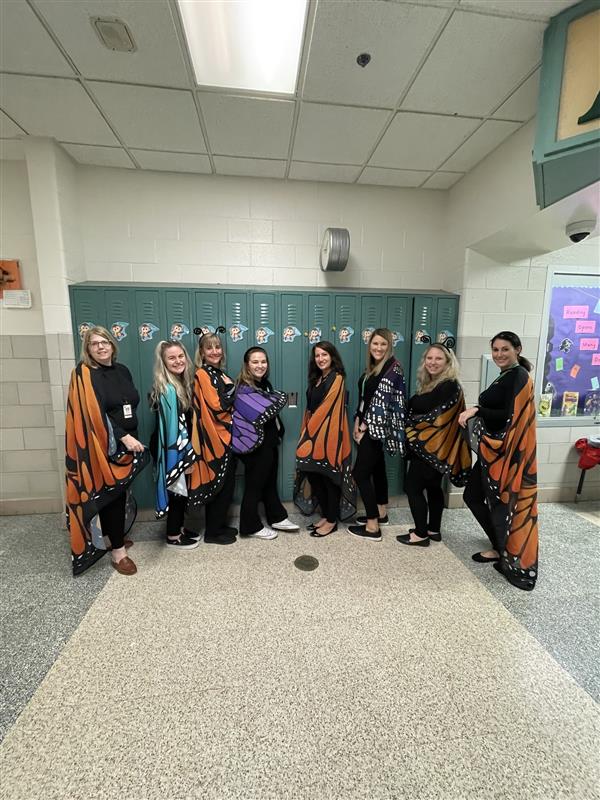 eight teachers pose in front of a bank of lockers - they are wearing colorful butterfly capes