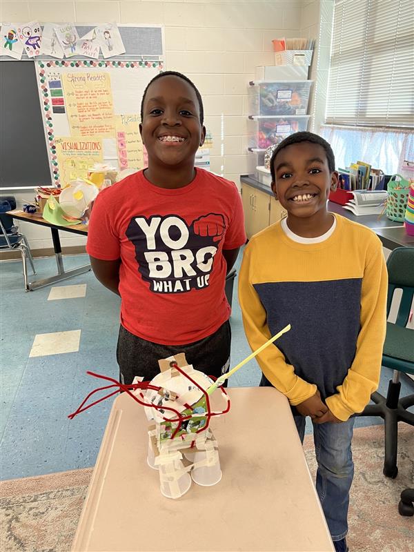 two student pose proudly in front of a project they've created - it appears to be a robot or small mechanical vehicle