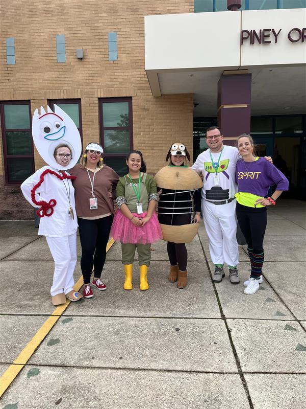 five teachers pose outside the entrance to Piney Orchard Elementary wearing their halloween costumes. One appears to be dressed as a spork, three are wearing 80s inspired outfits, one is dressed as the slinky dog from Toy Story, and one is a storm trooper from Star Wars