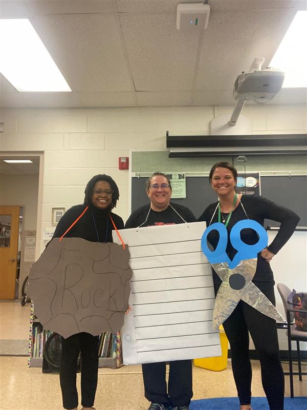 three teachers wear complementary Halloween costumes - one is a rock, one is scissors, and one is paper