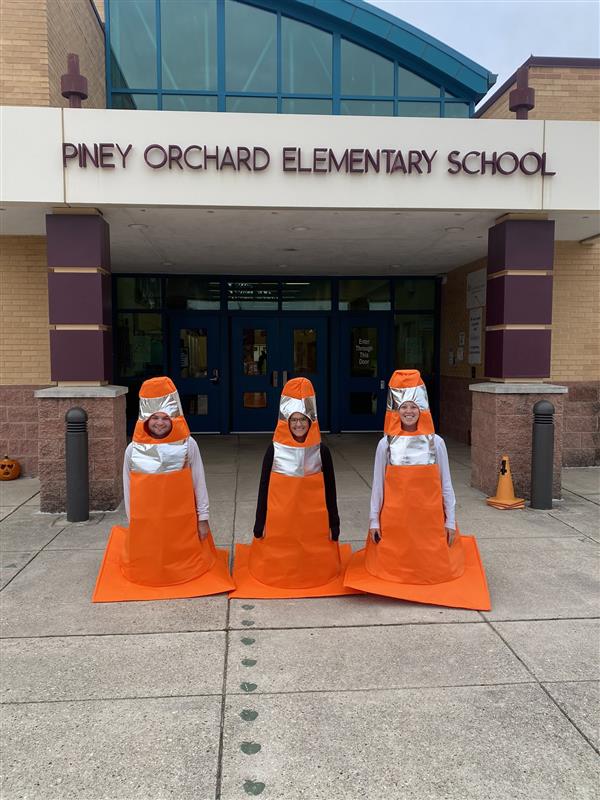 three faculty members are dressed as orange traffic cones and posed in front of the Piney Orchard Elementary entrance