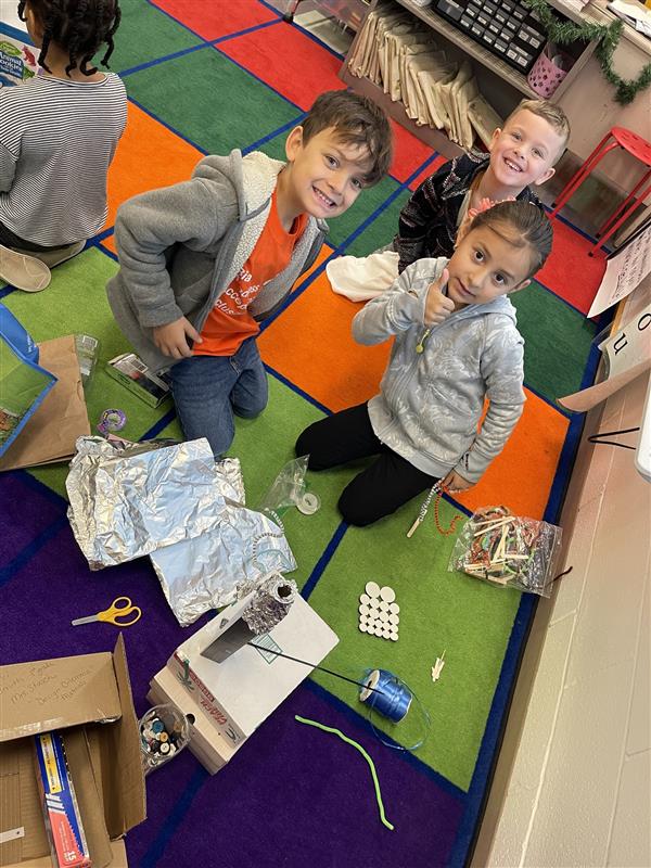 three students seated on a colorful carpet work together on a project involving foil, twine, and other supplies.