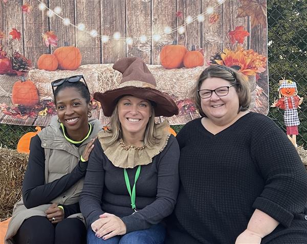 three teachers pose at a fall festival in front of a backdrop of hay bales and pumpkins 