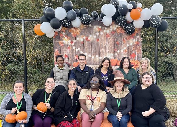 teachers pose together at a fall festival, in front of a balloon arch back drop. Some of the teachers are holding pumpkins, and one is dressed as a scarecrow