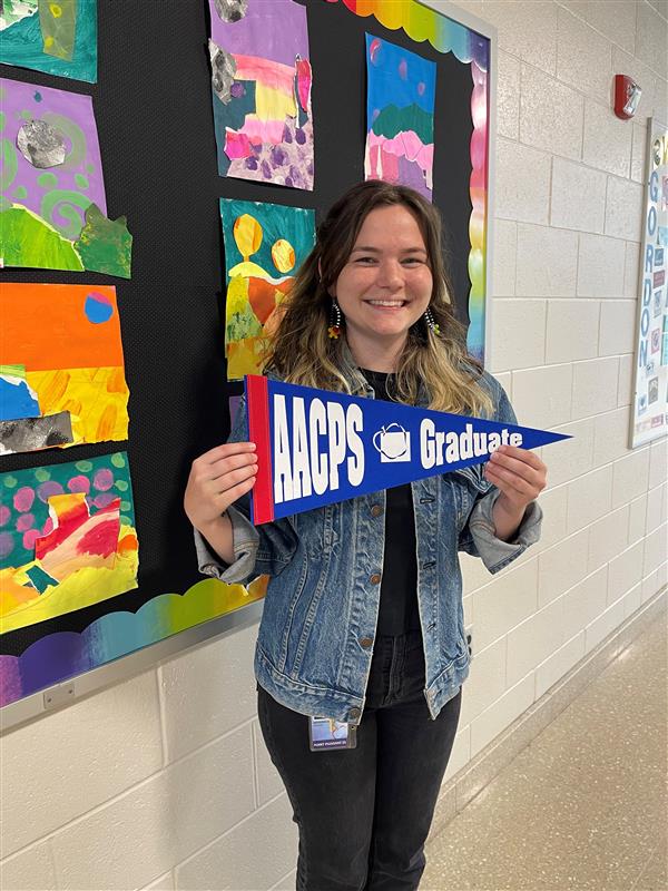 A student poses with a pennant reading "AACPS graduate". In the background are colorful pieces of art painted by students.