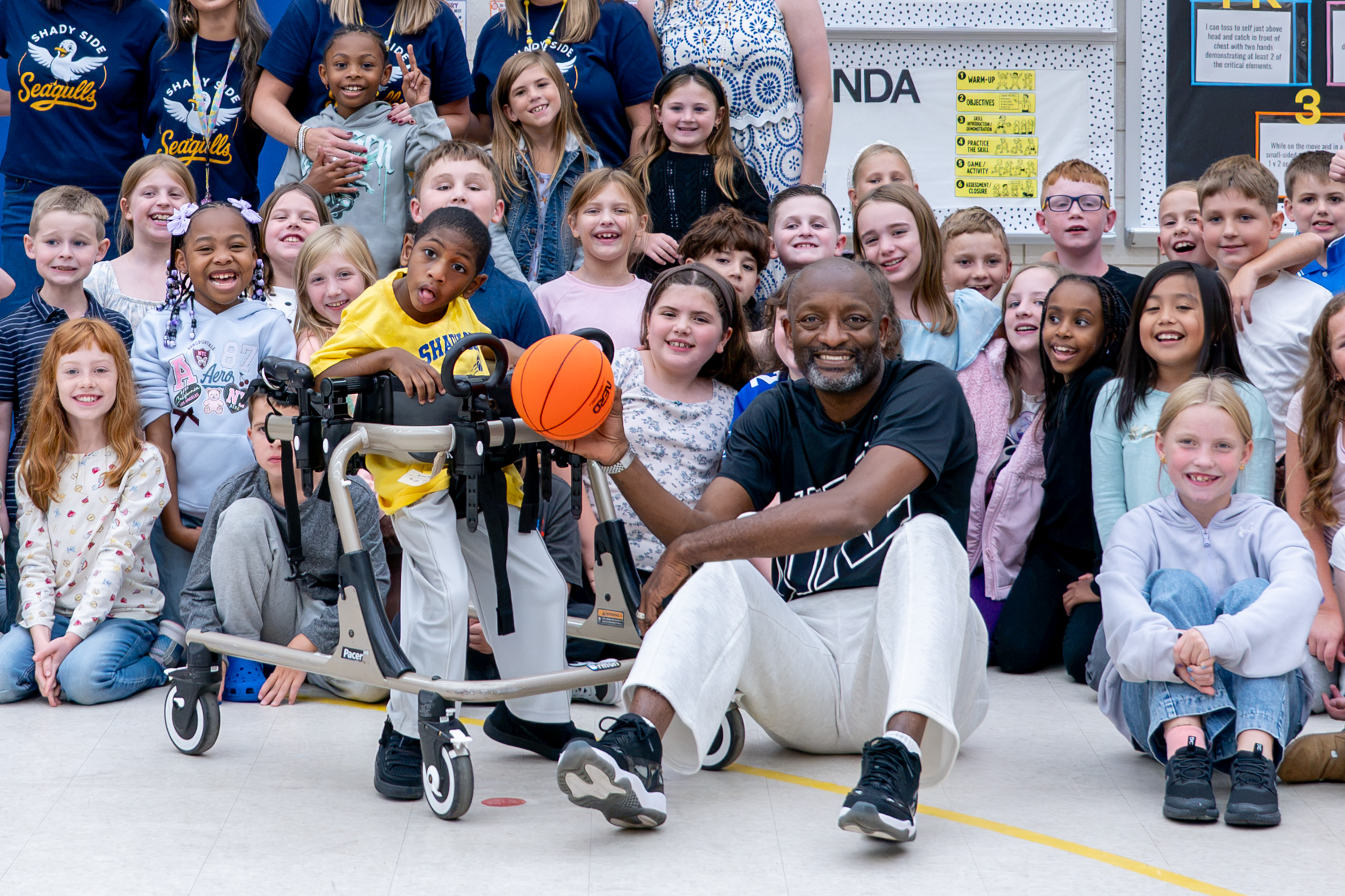 Man sits on floor holding basketball, child in wheelchair next to him, many children crouch or sit around them.