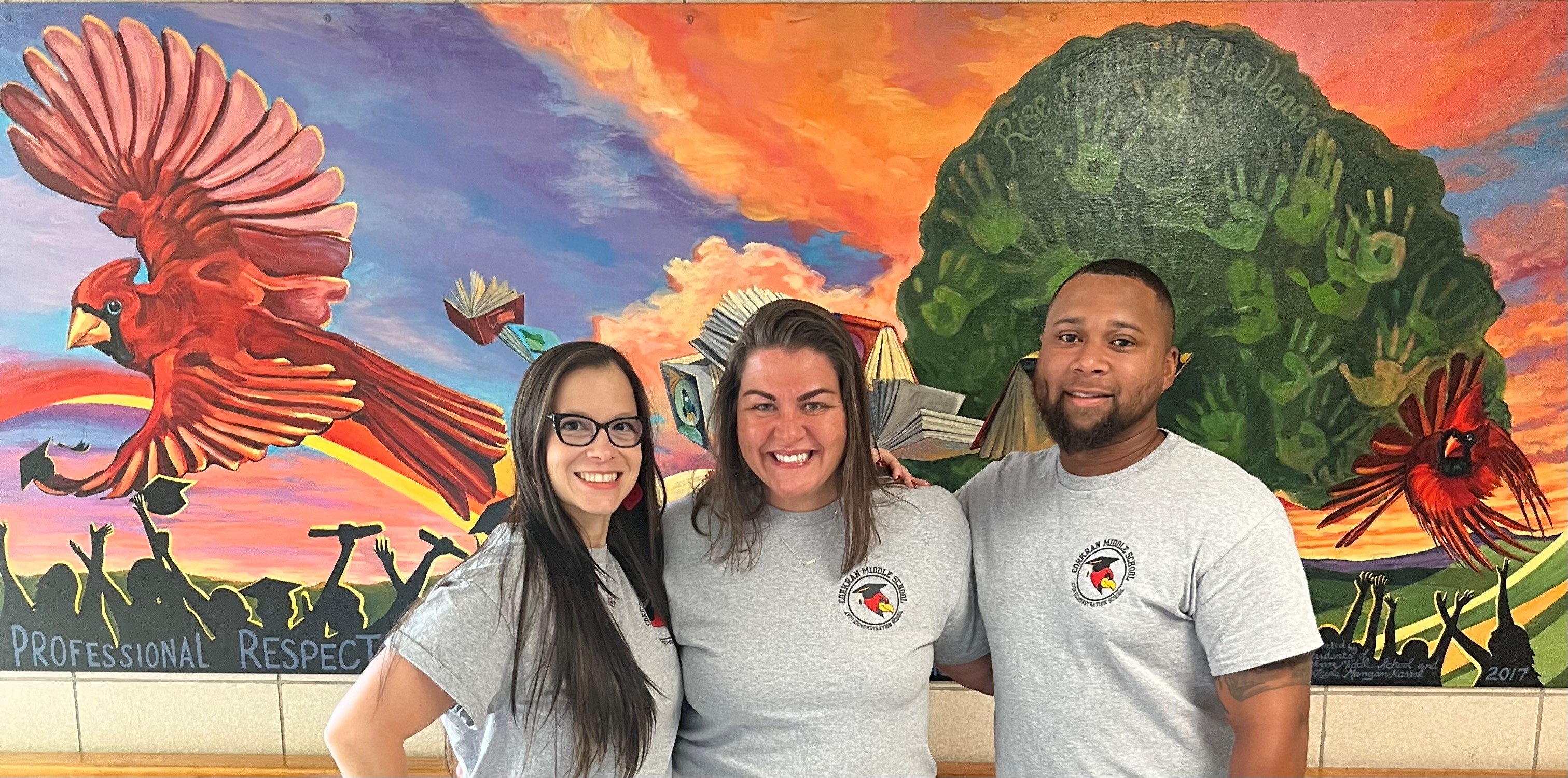Three people stand together in front of a mural with a bird, tree, and sunset.