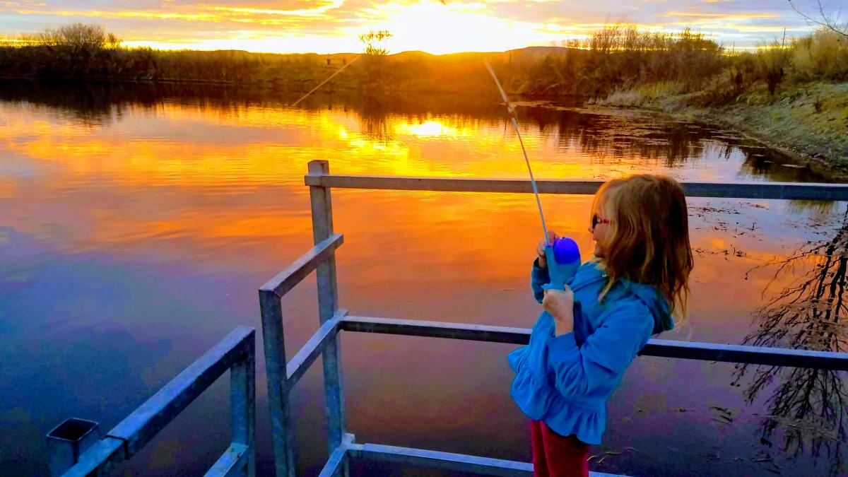 Child fishing on a lake at sunset, with the sun setting in the background.