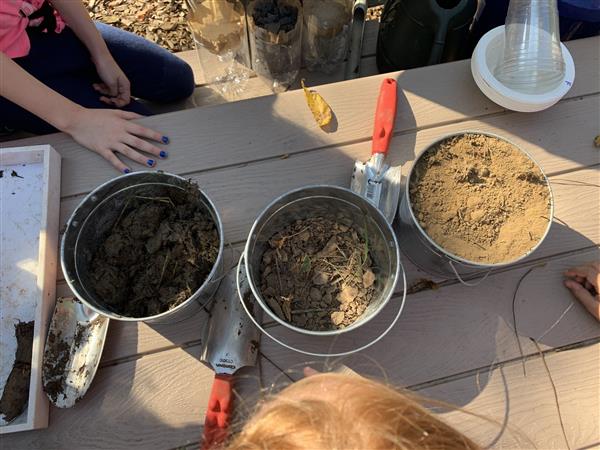 Children sitting on picnic table with buckets of dirt, enjoying outdoor activity.