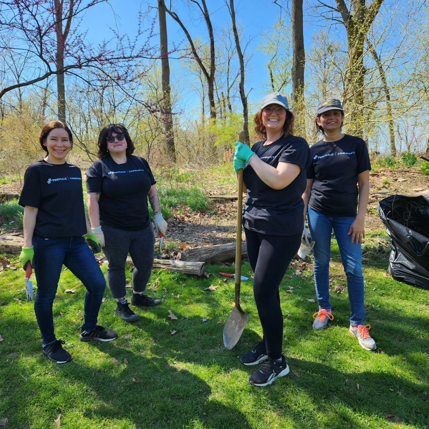 Four women in black shirts and pants standing in a park with shovels.