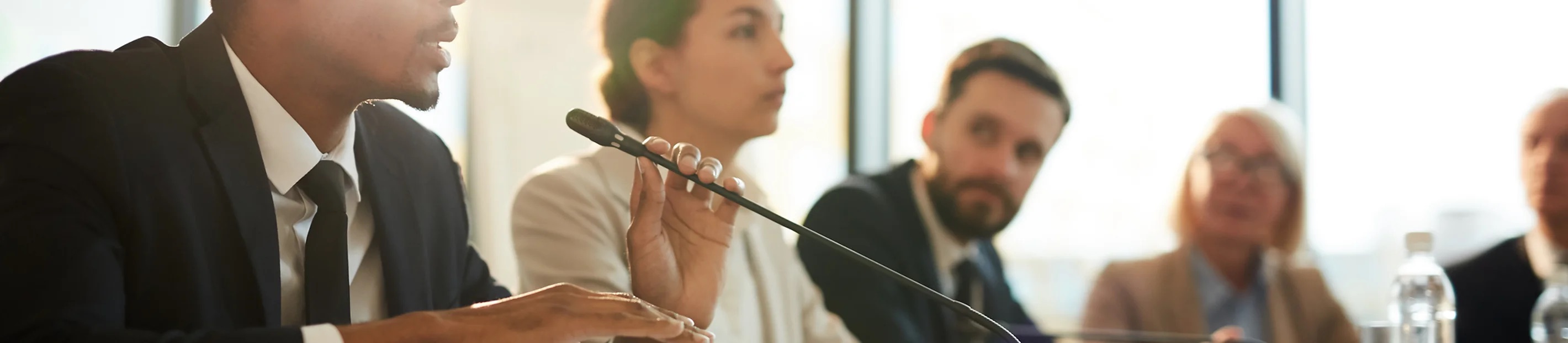 picture of the board in a meeting, a person is talking on the microphone and others are focused