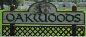 A wooden sign with the word "Oakwoods" written in white letters. The sign is mounted on a metal fence with a lattice design.