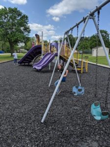 A playground with a girl playing swing with trees and blue sky at the background.