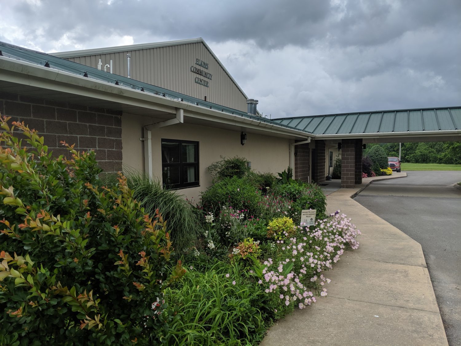 The image show the Elkins Community Center in Elkins, Arkansas. There are flowers planted in front of the building, and the text “ELONS” is visible on the awning. The Elkins Community Center is a large facility that offers a variety of programs and services to the community, including fitness classes, senior activities, and childcare.