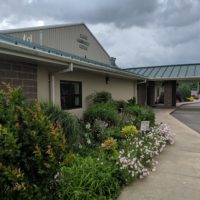 The image show the Elkins Community Center in Elkins, Arkansas. There are flowers planted in front of the building, and the text “ELONS” is visible on the awning. The Elkins Community Center is a large facility that offers a variety of programs and services to the community, including fitness classes, senior activities, and childcare.