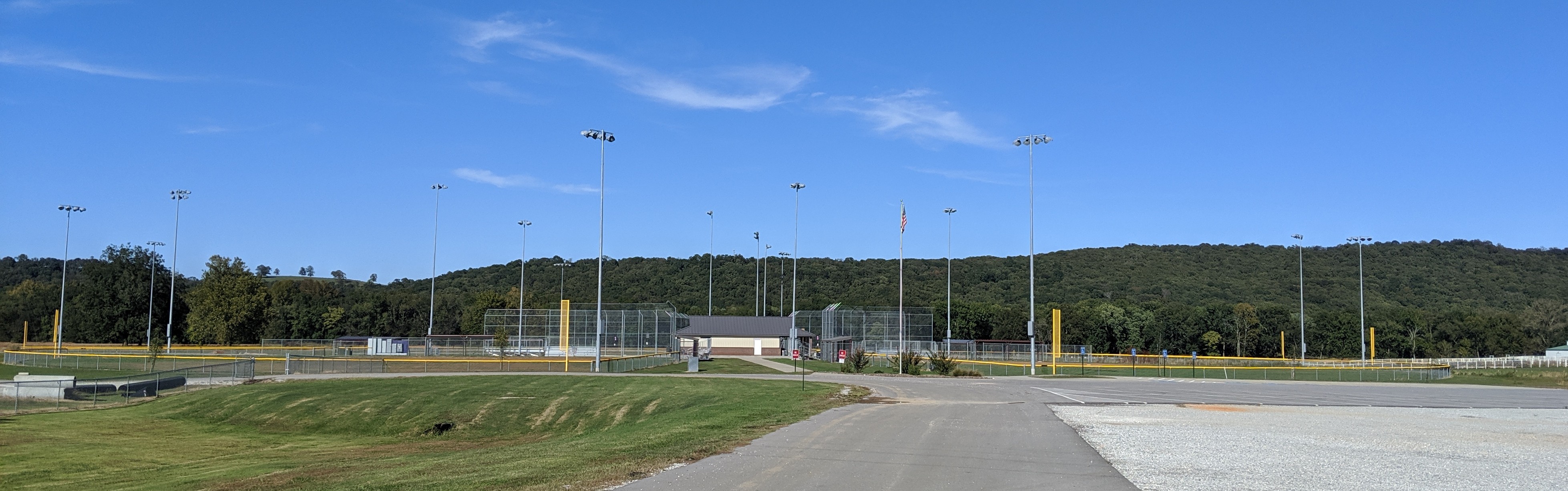 The image shows a dirt road leading to a baseball field with floodlights illuminating it. This scene suggests a night game is being played. There are trees lining the dirt road in the distance.