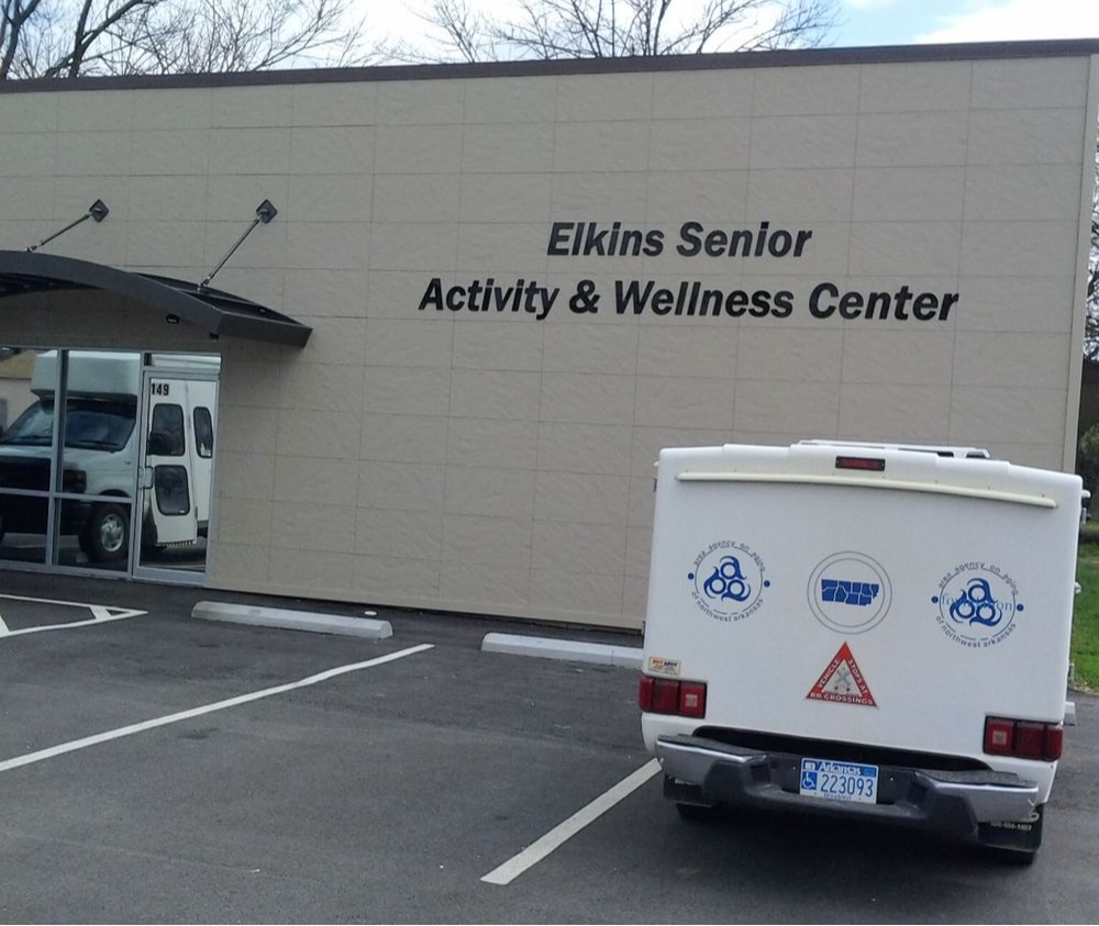 The image you sent me shows a white truck parked in front of a building with a sign that says "Elkins Senior Activity & Wellness Center" on it. The truck is a four-door pickup truck with a white camper shell. There are evergreen trees planted around the building.