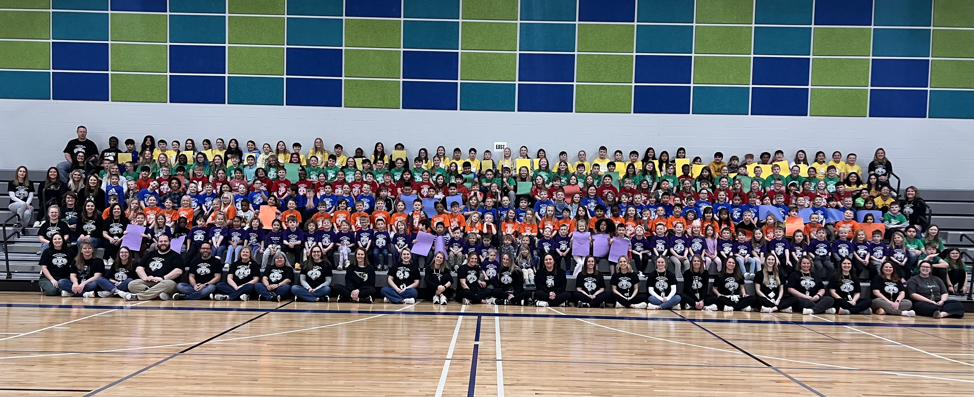 All school picture with grade level t-shirts of all colors.   K-5 students seated in the gym.