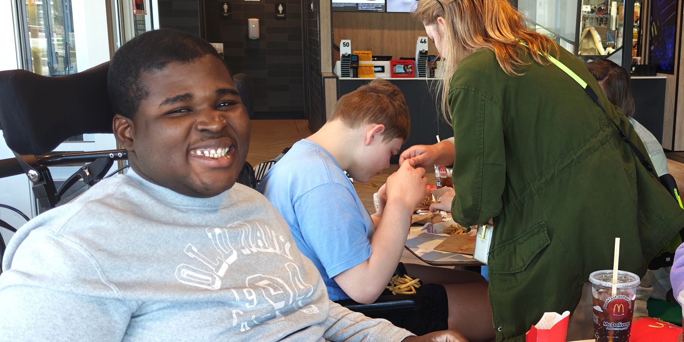 Three people sit around a table; one in a wheelchair smiles, another prepares food, and a third holds a drink.