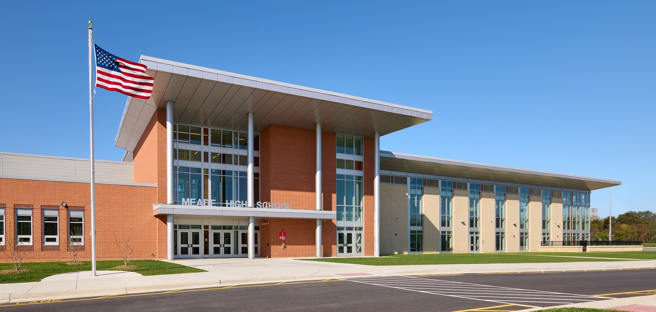 A modern building with a flagpole. It has many windows, a large entrance, and a paved road.