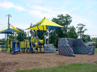 A colorful playground with yellow and blue equipment, a climbing wall, and shaded areas set against a backdrop of green trees and a clear sky.