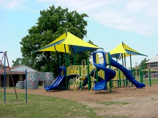 A colorful children’s playground with blue slides, yellow and green equipment, and yellow shade canopies, set against a backdrop of trees and a clear sky.