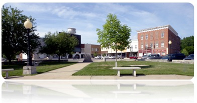 The image shows a panoramic view of Clinton Lake Park in Clinton, Illinois. The park has a variety of amenities including a walking path, a playground, and a bandshell. In the foreground of the photo, there is a bench and a large tree with a wide trunk and many branches. In the background, you can see the lake and some buildings.