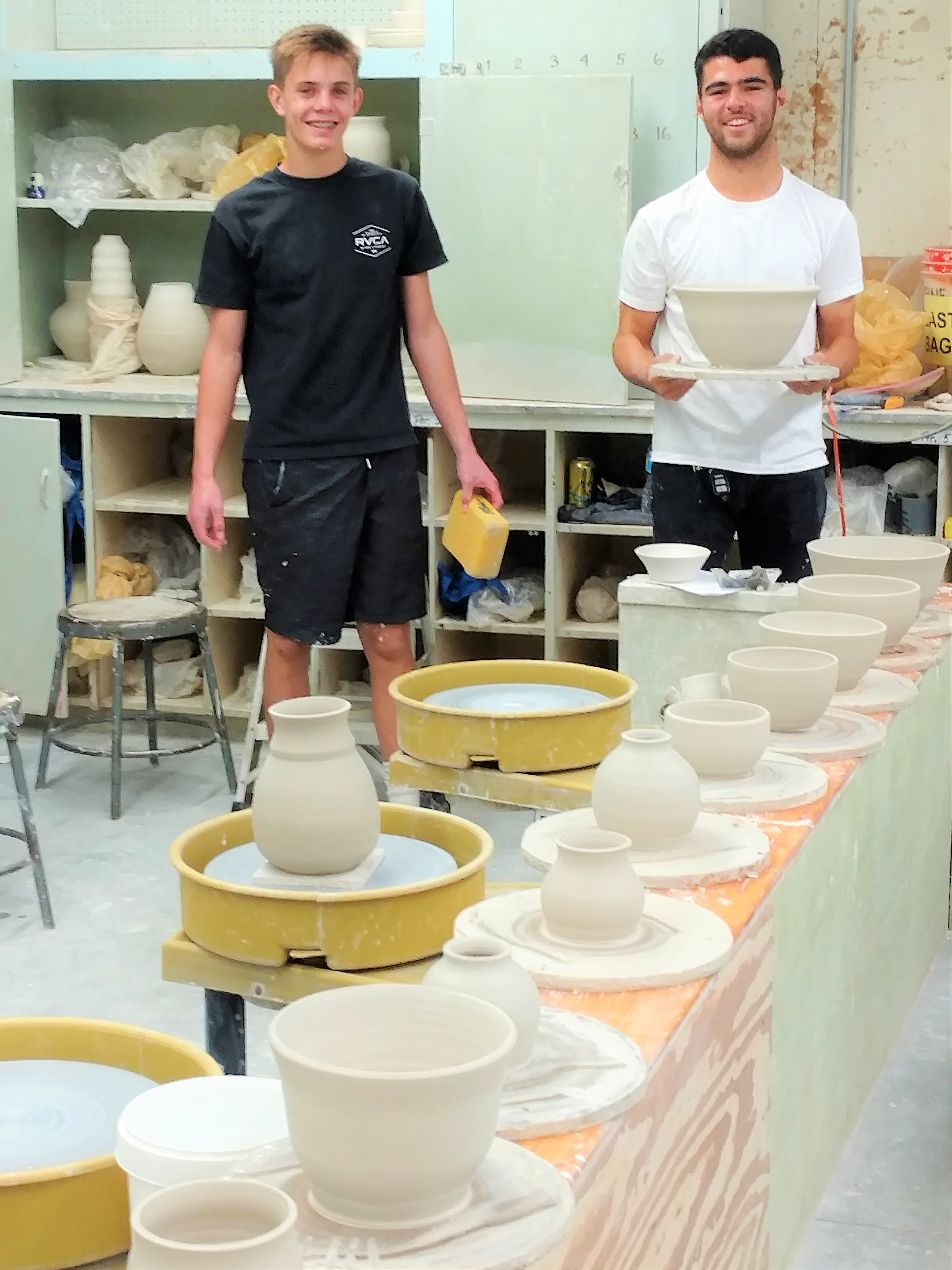 Two students smiling at the camera inside the ceramics classroom along with a row of ceramic plates and cups next to them.