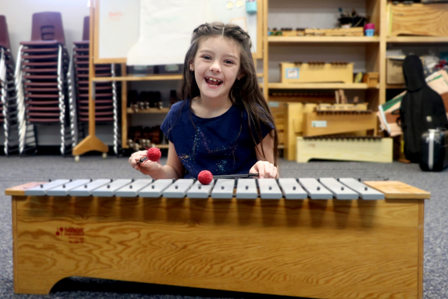 A young girl smiles brightly while playing a xylophone in a music classroom. 