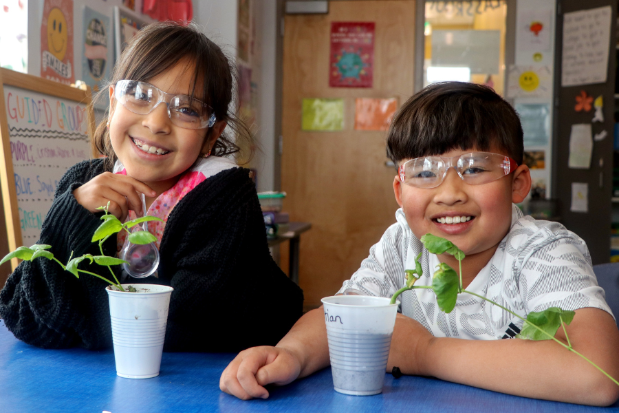 Two young students wearing safety goggles smile at the camera while sitting at a table. Each student has a small plant growing in a cup in front of them.