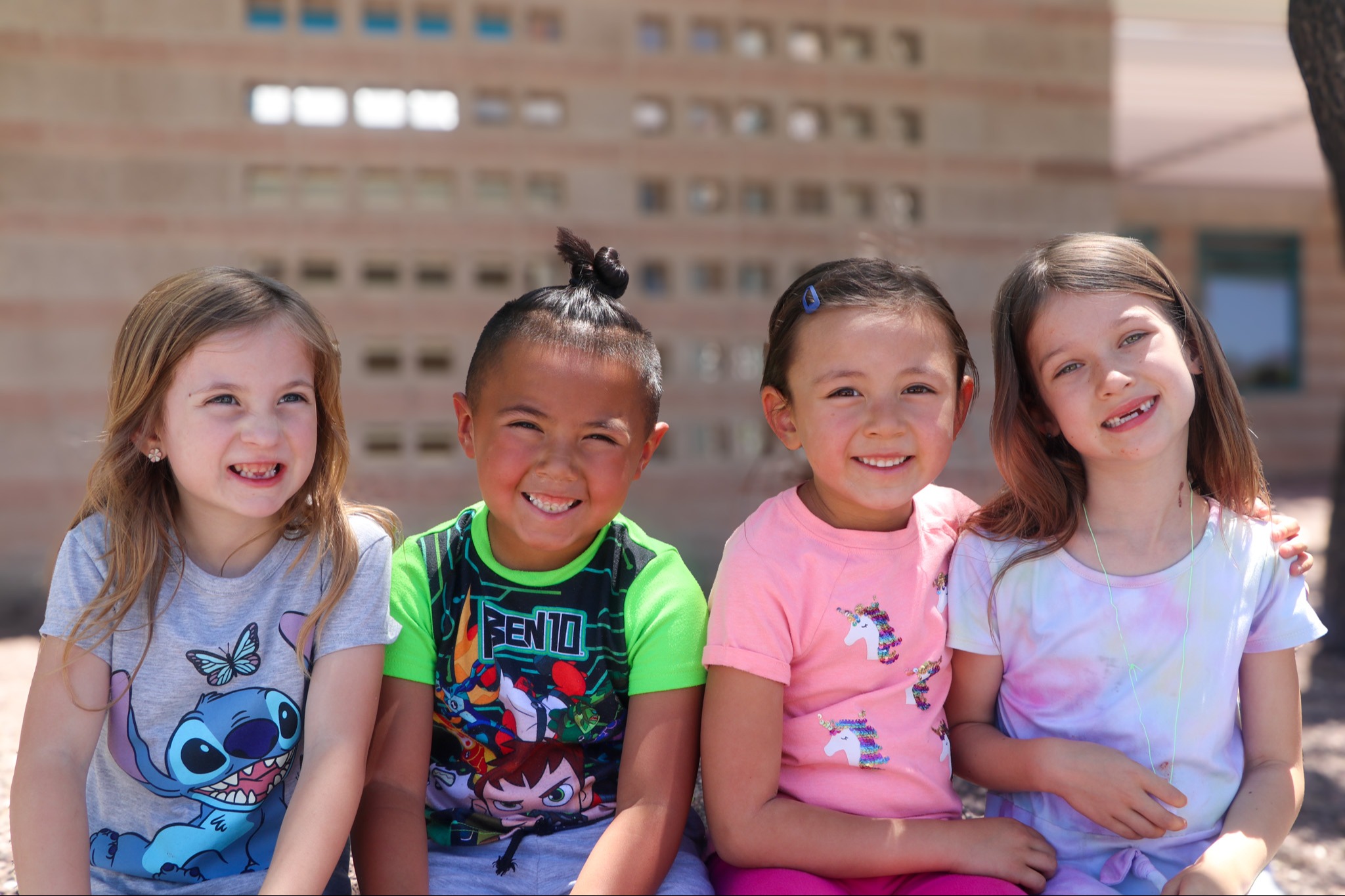 A group of four smiling children sitting together outdoors, wearing colorful outfits. 