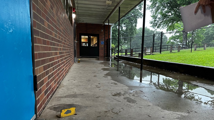 Covered walkway with flooded water, a brick wall to the left, double black doors in front, and green grass to the right. 