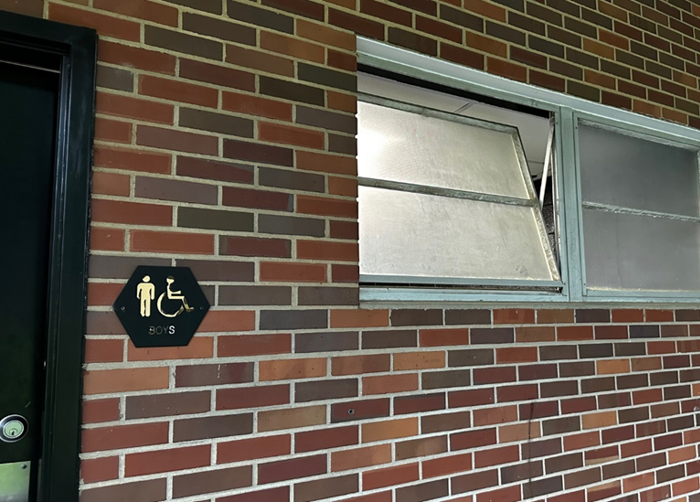 An exterior brick wall with windows at the top cracked open. The doorway has an restroom sign. 