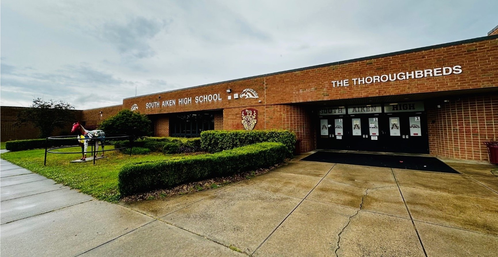 Front of a brick school building with a green space in front and a statue of a horse. 