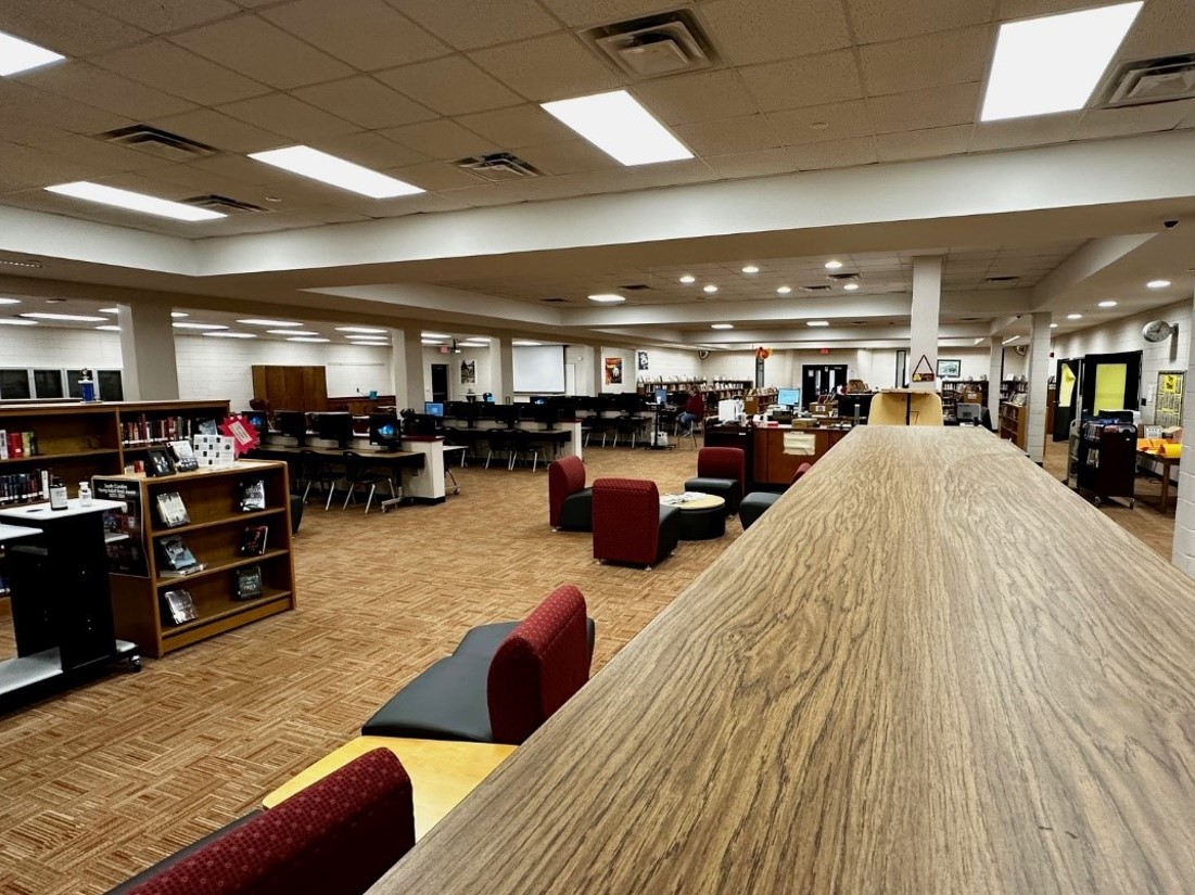 Large room that resembles a student library with long tables, books and colorful seating in maroon, black and yellow. 