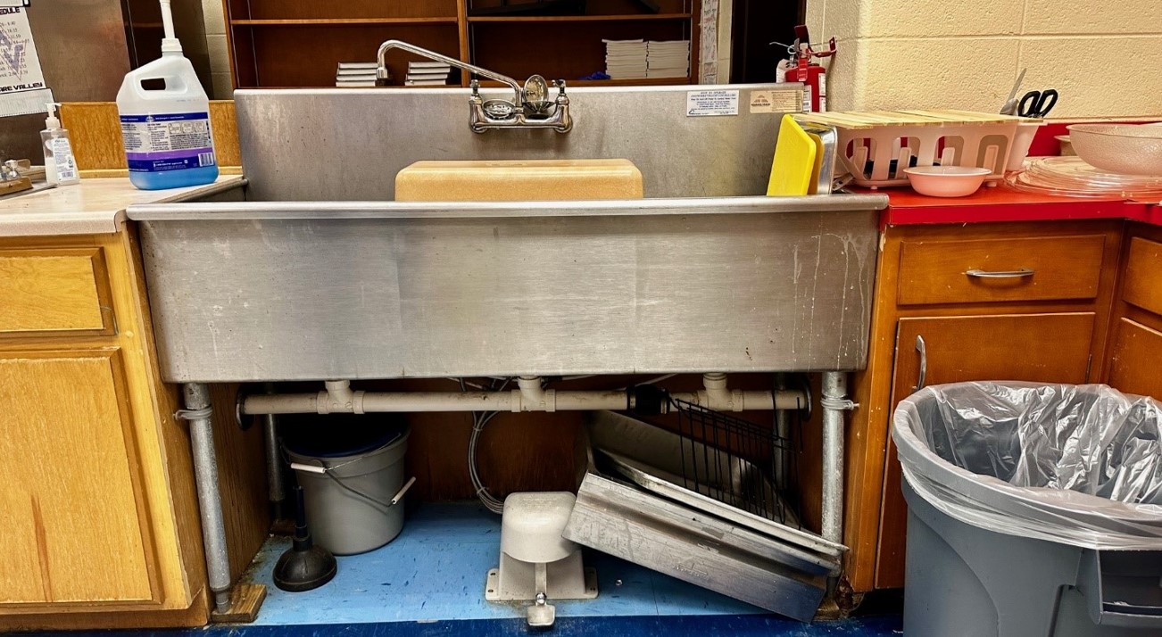 A sterling sink in a wooden cabinet with lots of pots, pans and clutter. 