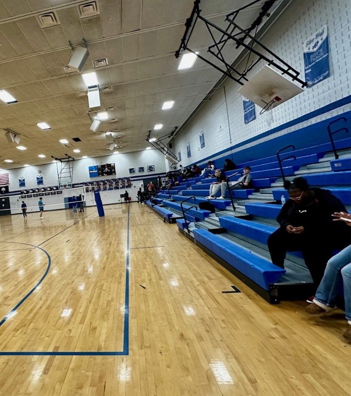 Indoor basketball gym with a few people sitting on blue bleachers. 