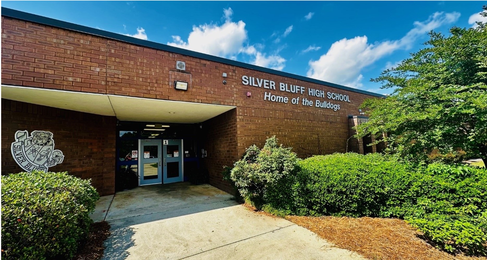 Exterior entrance to a one-level school. Bushes are on either side of the cement walkway to the double doors painted baby blue. 