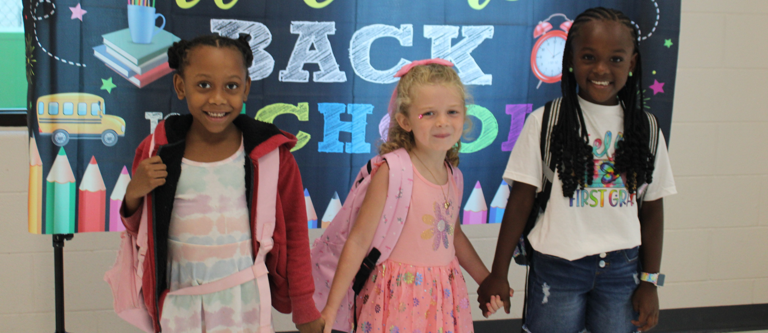Three elementary students holding hands in front of Welcome Back to School Banner.