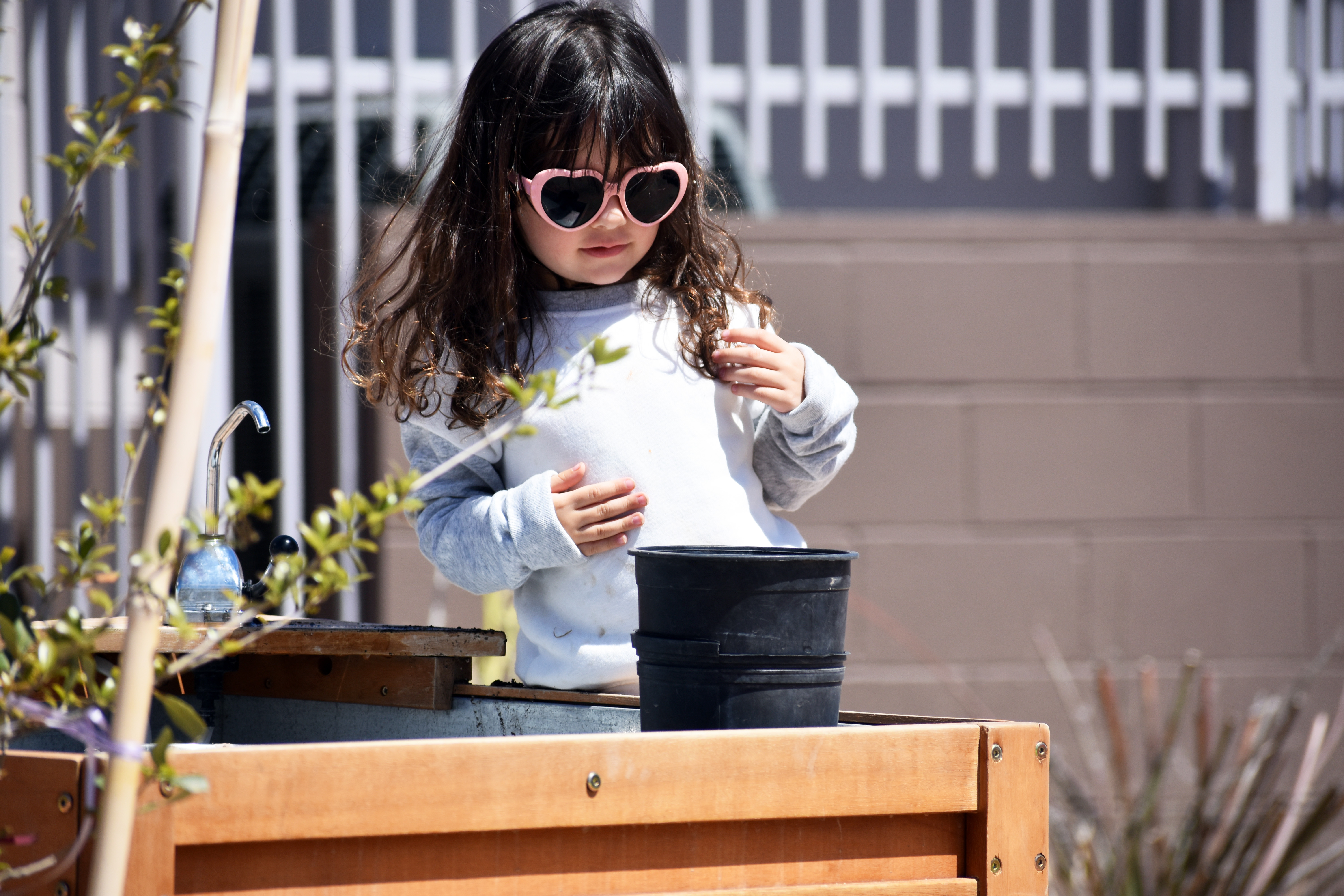 A young girl with long dark hair, pink heart-shaped sunglasses, and a light gray sweater stands by a wooden planter box, holding a small black pot.