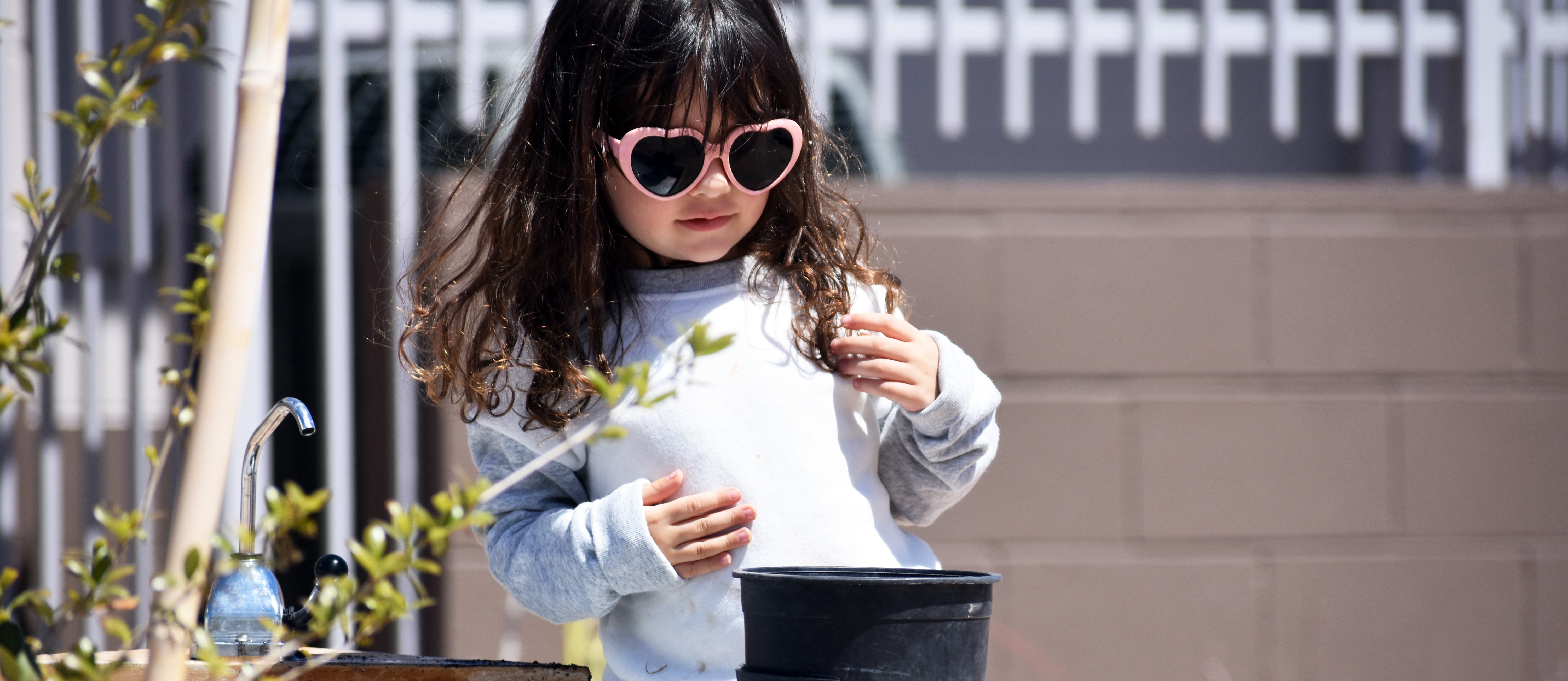 A young girl with long dark hair, pink heart-shaped sunglasses, and a light gray sweater stands by a wooden planter box, holding a small black pot.