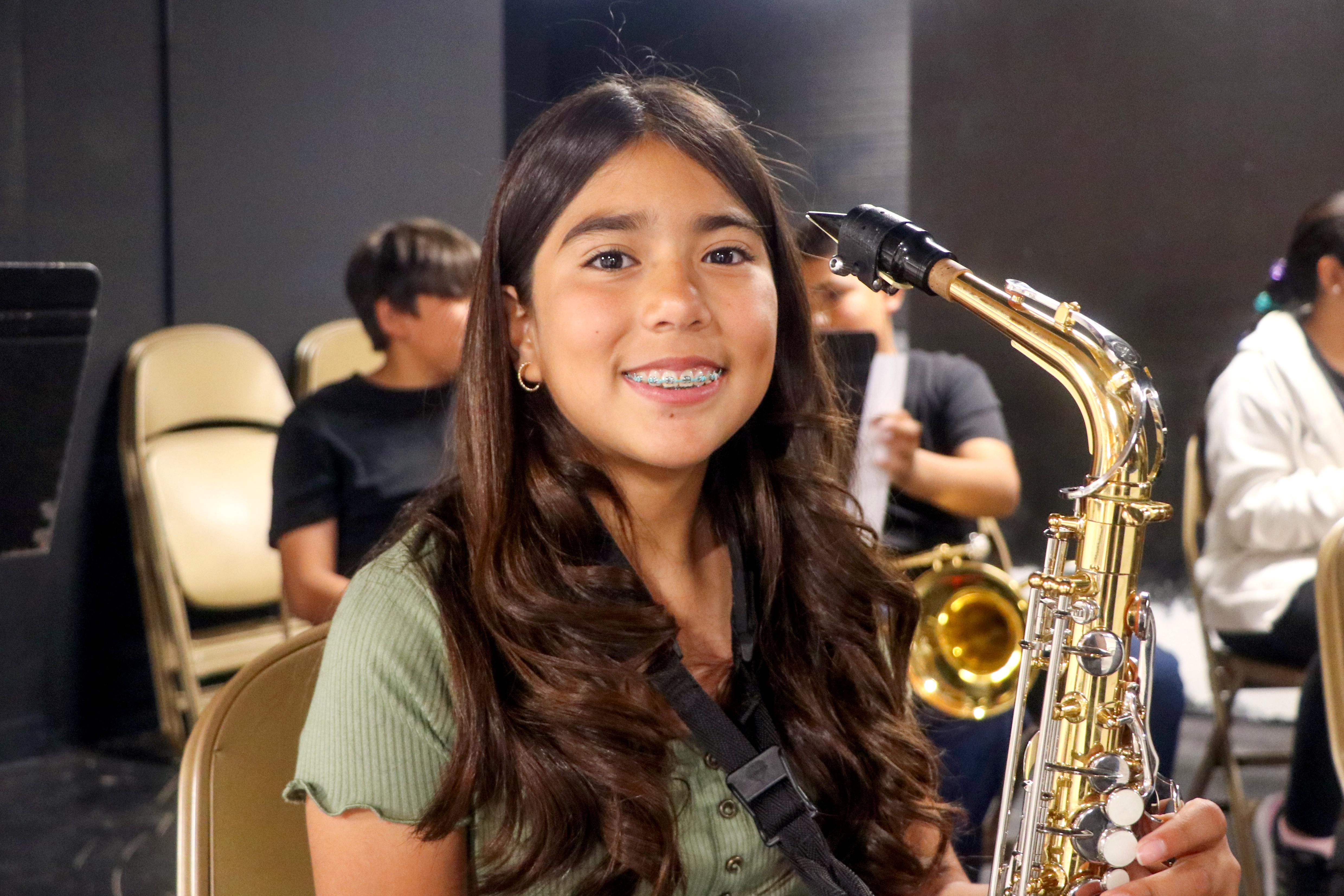 A girl sits, smiling and holding her saxophone. 
