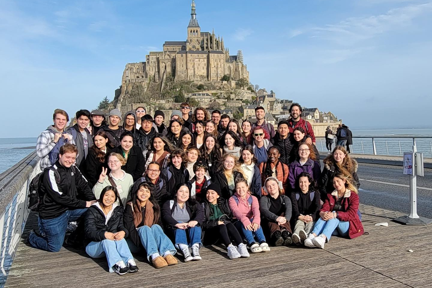 A large group of students pose for a photo on a pier with Mont Saint-Michel in the background.