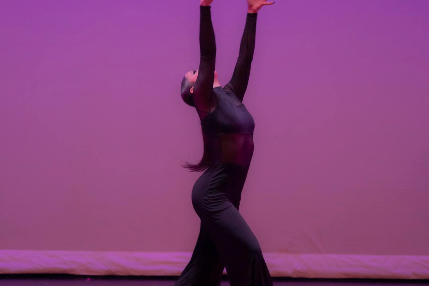 A dancer in a black outfit striking a dramatic pose with arms extended upwards against a purple-lit background on stage.
