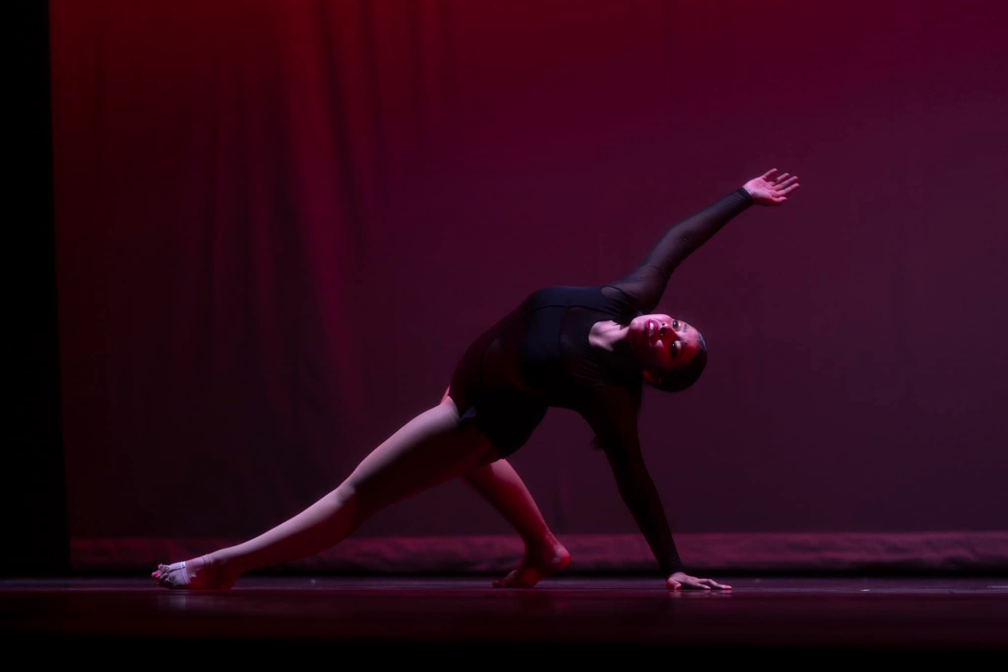 A dancer in a black outfit striking a dramatic pose with arms extended upwards against a purple-lit background on stage.