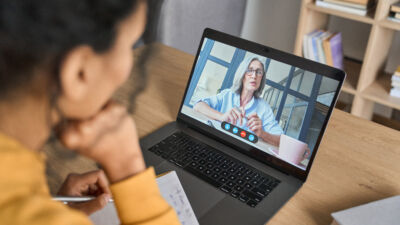 A woman sits at a desk taking notes while participating in a video call on a laptop. On the screen, another woman with glasses speaks from a modern office space. Books and papers are visible in the background, suggesting a home or office setting.