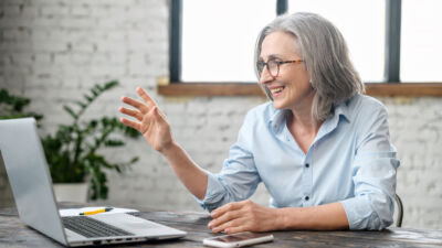 An older woman with gray hair and glasses smiles and waves while participating in a video call on a laptop. She is seated at a desk in a bright, modern room with a white brick wall and large windows in the background. A smartphone and notepad lie on the table in front of her.