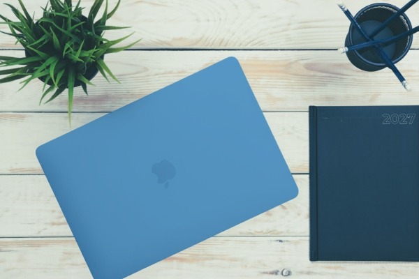 Indigo MacBook Neo sits closed on white wooden desk with green houseplant, blue journal and pencil cup with pencils in it.