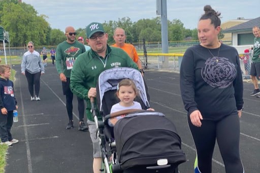 Photo of a child being pushed by his father on a toddler cart alongside other runners