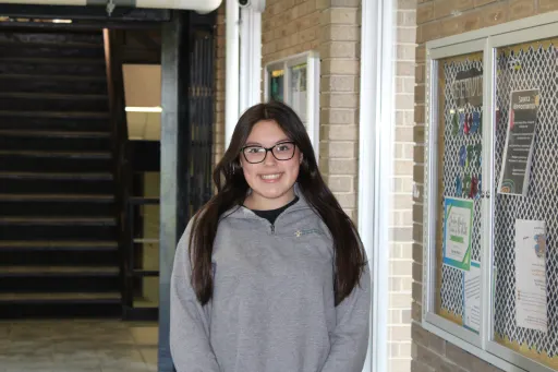 A student wearing a grey hoodie stands in a well-lit indoor hallway